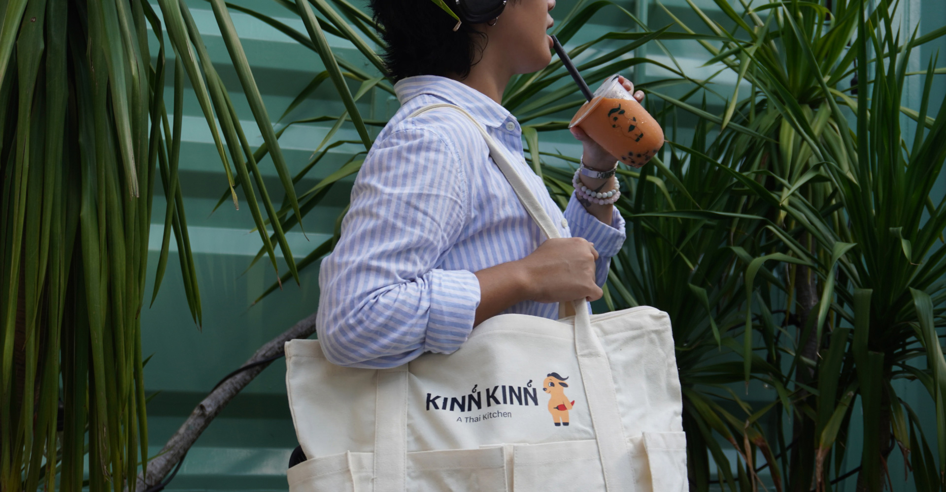 A photo of a person with the Kinn Kinn merch tote bag on their shoulder holding a drink with Kinn Kinn's mascot printed on it